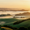 A sunrise over a valley with fog and hills logo